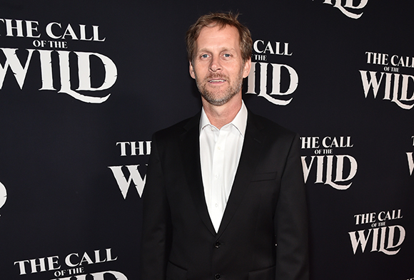 A man with light hair and a beard wearing a black suit and white shirt stands before a movie backdrop.