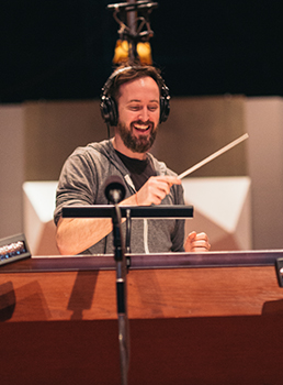 A smiling man wearing headphones conducts with a baton behind a music stand.