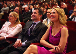 A man in a suit and a woman in a magenta dress smile while sitting in a crowded theater.