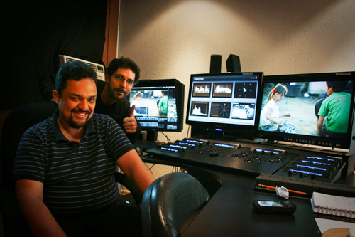 Two men sit in a dark studio in front of three computer monitors displaying video editing software.