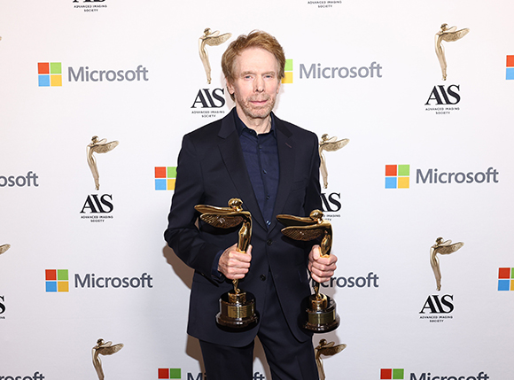 A man in a dark suit holds two gold winged trophies in front of a white backdrop with Microsoft and AIS logos.