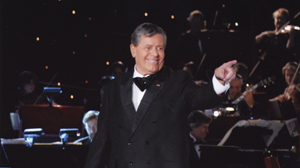 A smiling man in a black tuxedo and bowtie points toward the camera in front of an orchestra.