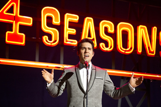 A man in a grey suit sings into a microphone before a large orange neon sign that reads '4 SEASONS'.