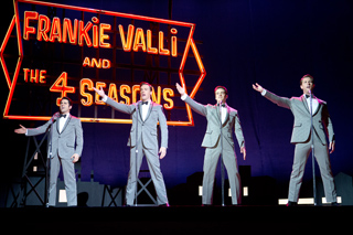 Four male singers in grey suits perform on stage in front of a large neon Frankie Valli and the Four Seasons sign.