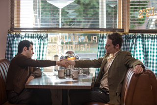 Two men in jackets sit at a diner booth and shake hands across a table.