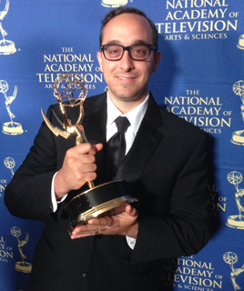 A man in a black suit and glasses holds a gold trophy against a blue background with white text.