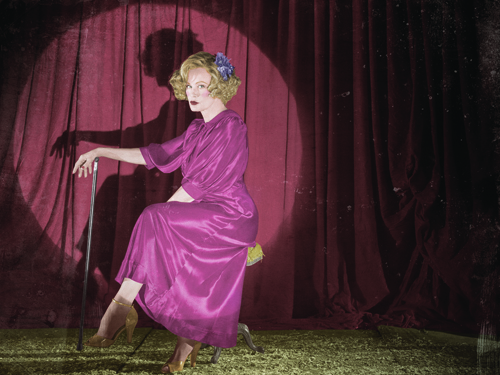 A woman in a pink satin dress holds a cane while posing in front of red velvet curtains.