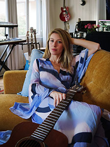 A woman in a blue patterned dress sits on a yellow sofa holding an acoustic guitar.