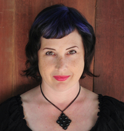A woman with dark, wavy hair with purple streaks and freckles smiles at the camera. She wears a black necklace with a dark pe