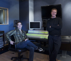 Two men posing in a recording studio with a mixing console and monitors.