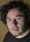 Close-up portrait of a man with dark curly hair and blue eyes looking toward the camera.