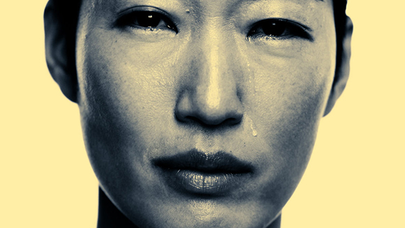 Close-up of a person's face with tears streaming down their cheek against a yellow background.