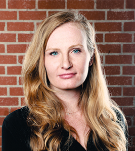 A woman with long blonde hair and blue eyes looks directly at the camera against a red brick background.