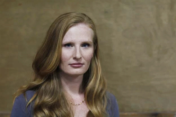 A woman with long, wavy reddish-blonde hair and blue eyes looks forward against a neutral background.