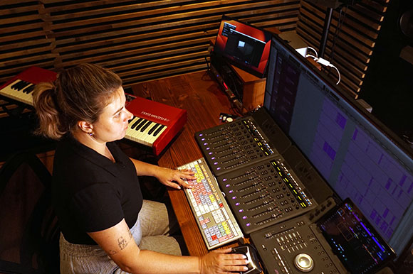 A woman sits at a desk with a computer monitor, mixing console, and MIDI keyboard in a studio.