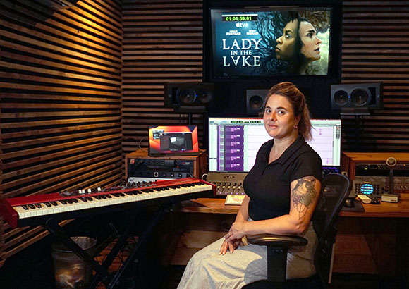 A woman sits in a music studio with a keyboard, monitors, and a screen displaying Lady in the Lake.