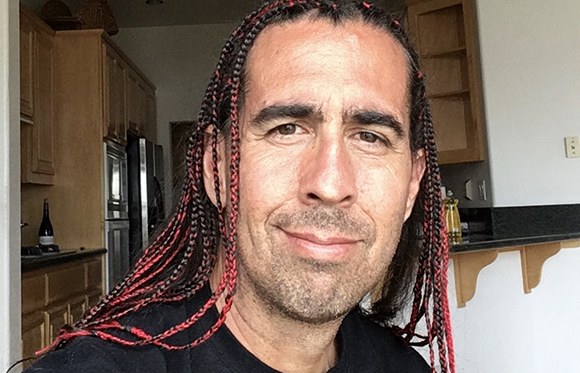A man with long, thin red and black braids smiling in a kitchen.