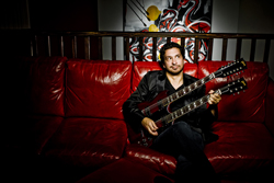 A man in a black shirt sits on a red leather couch holding a double-neck electric guitar.
