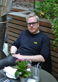 A man with glasses and a black long-sleeve shirt sits at a table with a small potted plant.