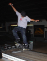 A person in a grey shirt and dark jeans performs a trick on a skateboard on an indoor ramp.