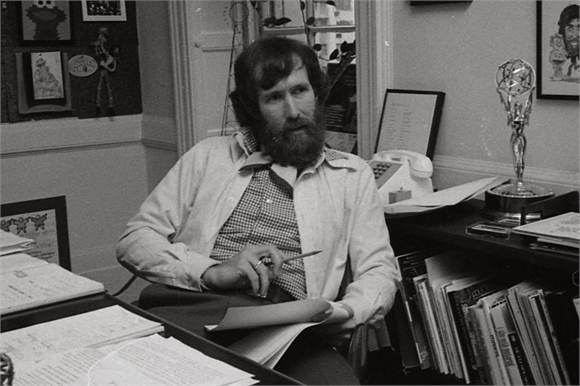 A bearded man in a button-down shirt sits at a desk covered in papers, books, and a trophy.