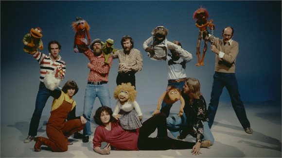 A group of people in casual clothing posing with several large hand puppets against a blue background.