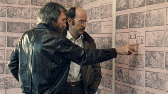 Two men stand before a wall covered in storyboard sketches, one pointing at a specific panel.