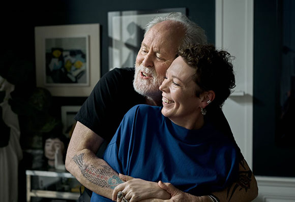 A man with a white beard hugs a smiling woman with short curly hair from behind.
