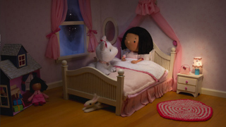 A doll with black hair sits in a small white bed next to a white stuffed animal in a pink room.