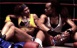 Two female boxers sit on the floor of a boxing ring, holding water bottles.