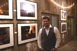 A man in a white shirt and grey vest stands before a wooden wall displaying framed photographs.