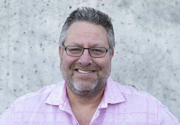 A smiling man with short salt-and-pepper hair and glasses wearing a pink patterned shirt.