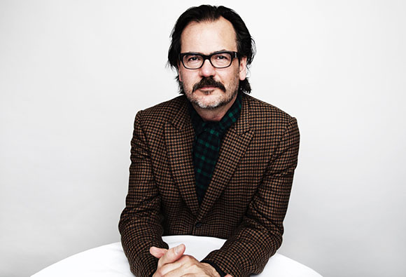 A man with dark hair, glasses, and a mustache wearing a brown checkered blazer and dark patterned shirt.