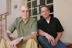 Two men sitting side by side on a ledge in front of a building with glass doors.