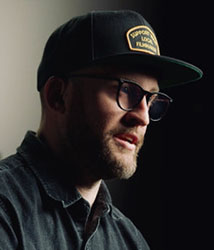 Side profile of a bearded man wearing a dark baseball cap and glasses against a dark background.
