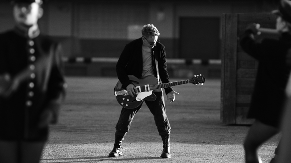 A man in a dark jacket and trousers plays an electric guitar while standing in an outdoor area.