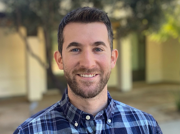 A smiling man with short dark hair and a beard wearing a blue and white plaid button-down shirt.