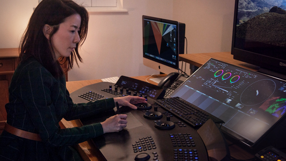 A woman uses a large control surface and multiple monitors in a dark post-production studio.