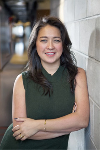 A smiling woman with long dark hair wearing a dark green sleeveless top, leaning against a light gray brick wall.