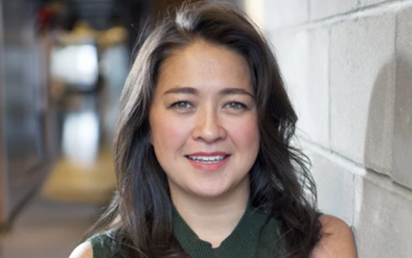 A smiling woman with long dark hair leans against a light gray cinder block wall in a hallway.