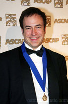 A smiling man in a black tuxedo and bowtie wearing a blue ribbon with a gold medal.