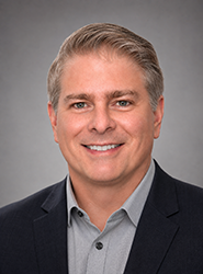 A smiling man with light brown hair wearing a grey shirt and a dark navy blazer against a grey background.