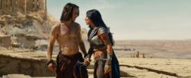 A shirtless man and a woman in armor stand together in a vast, sandy desert landscape.