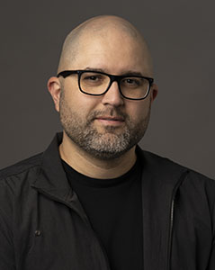 A bald man with glasses and a salt-and-pepper beard wearing a black jacket.