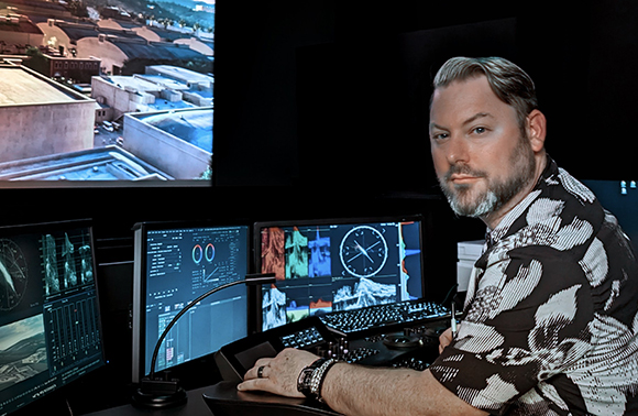 A man in a patterned shirt sits at a desk with several computer monitors displaying data and maps.