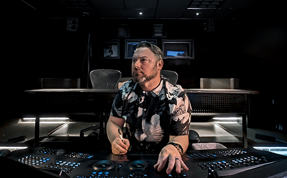 A man in a patterned shirt sits at a large video editing control surface in a dark studio.