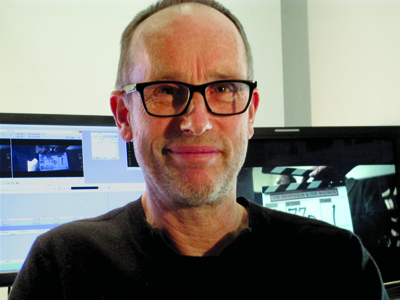A man with short hair and black-rimmed glasses smiling in front of computer screens.
