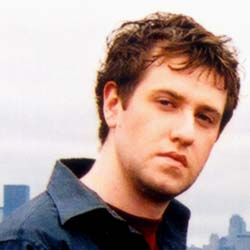 A young man with short brown hair and a dark shirt looks directly at the camera against a pale sky.