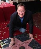 A man in a dark blazer kneels on a red carpet next to a star on the Hollywood Walk of Fame.