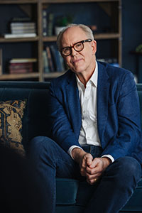 A man with glasses wearing a blue blazer and white shirt sits on a dark velvet sofa.
